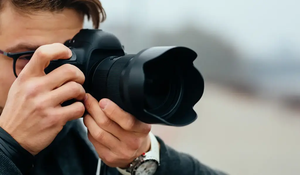 Iemand die een professionele camera dicht bij zijn gezicht houdt en scherpstelt op een opname in een buitenomgeving. De scène straalt concentratie en creativiteit uit.