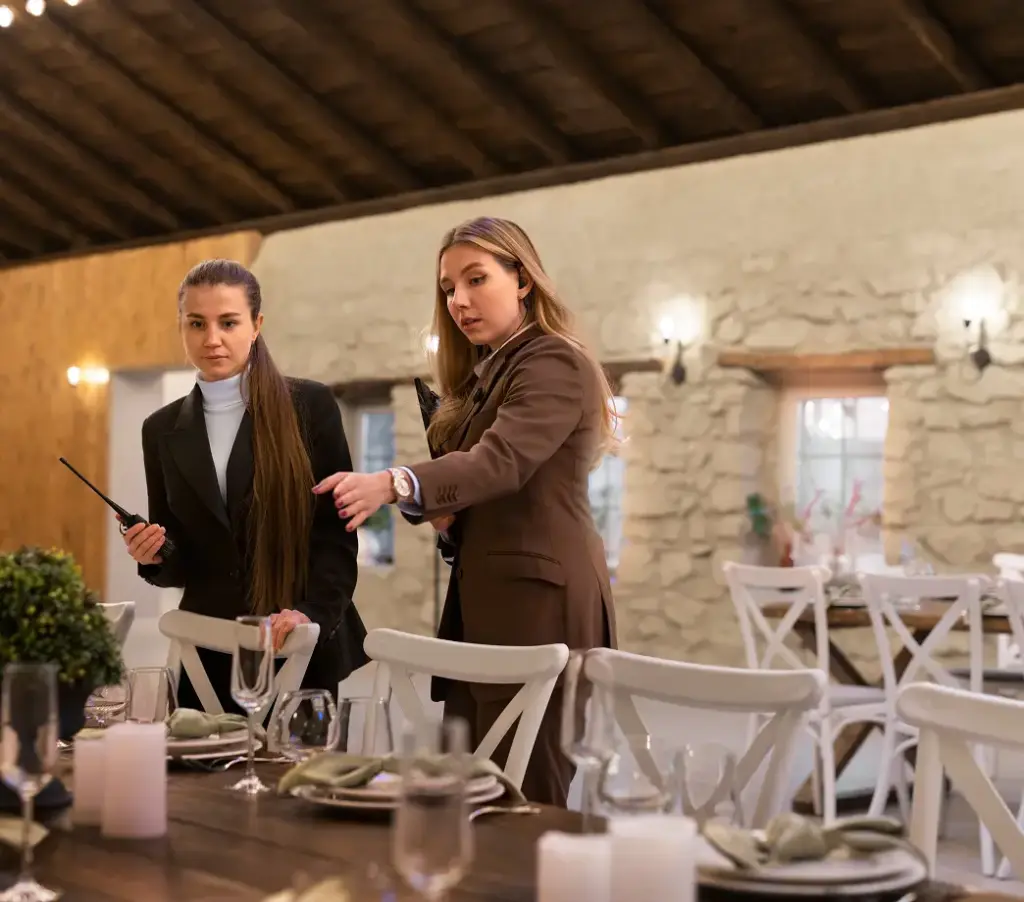 Twee vrouwen in formele kleding dekken een rustieke eettafel in een elegante, sfeervol verlichte kamer. Houten balken en stenen muren zorgen voor een gezellige sfeer.