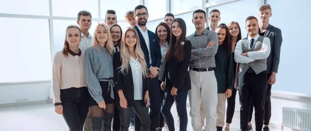 Een groep van 16 jonge professionals staat in een helder verlicht kantoor en glimlacht zelfverzekerd. Ze zijn gekleed in business-casual kleding, wat een gevoel van teamgeest en positiviteit uitstraalt.