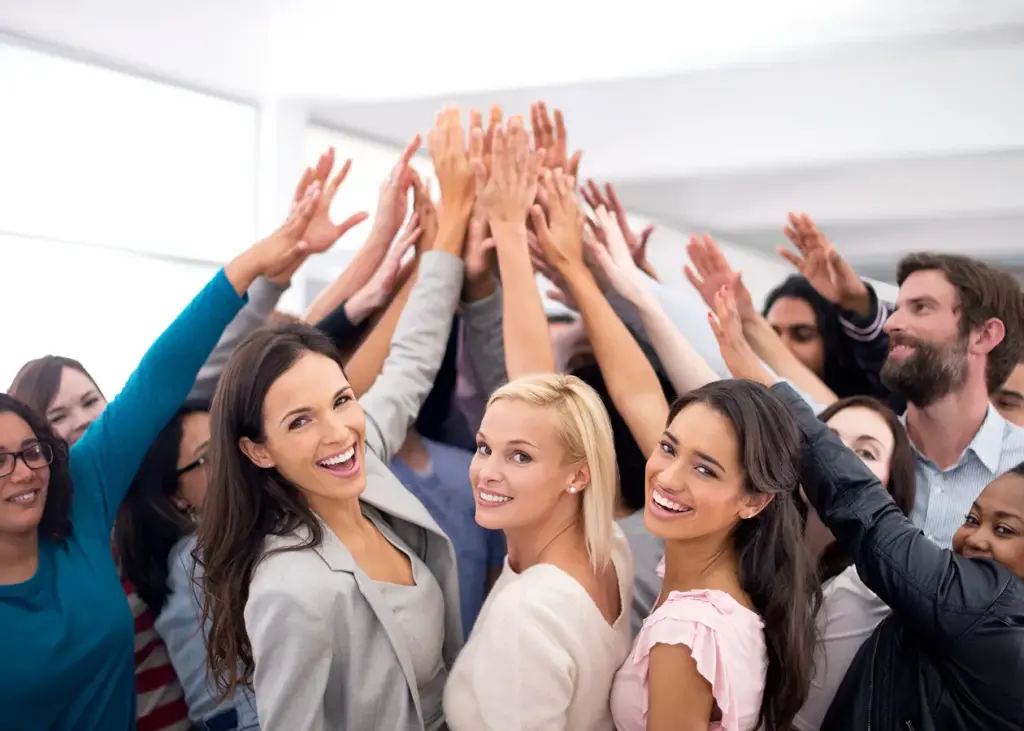 Een diverse groep mensen die glimlacht en tegelijkertijd de hand opsteekt in een lichte ruimte, waardoor een gevoel van teamgeest, saamhorigheid en positieve energie wordt uitgestraald.